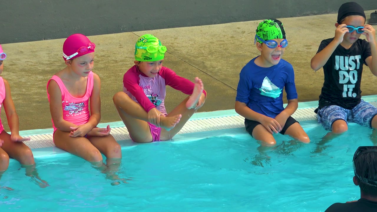 Children in swim caps sit beside turquoise pool water while one child adjusts goggles.