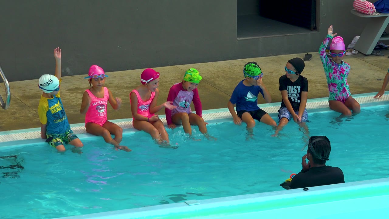 In Cabo San Lucas, Mexico, children wearing swim caps and goggles sit along a pool edge while an adult swim instructor faces them from the foreground water.