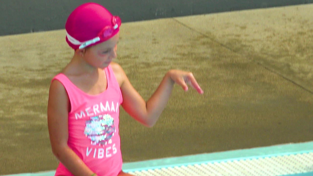 A girl in a pink swimsuit and pink swim cap sits at the pool edge in Cabo San Lucas, Mexico, with sunlit deck and blue water below.