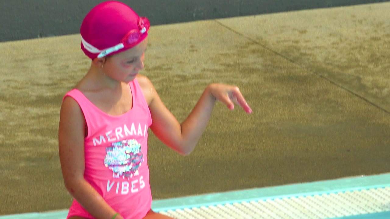Girl in pink swim cap gestures beside the pool.