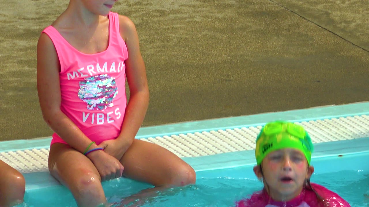 A seated girl in a pink swimsuit watches from the pool edge in Cabo San Lucas, Mexico, as a child in a green swim cap surfaces nearby.