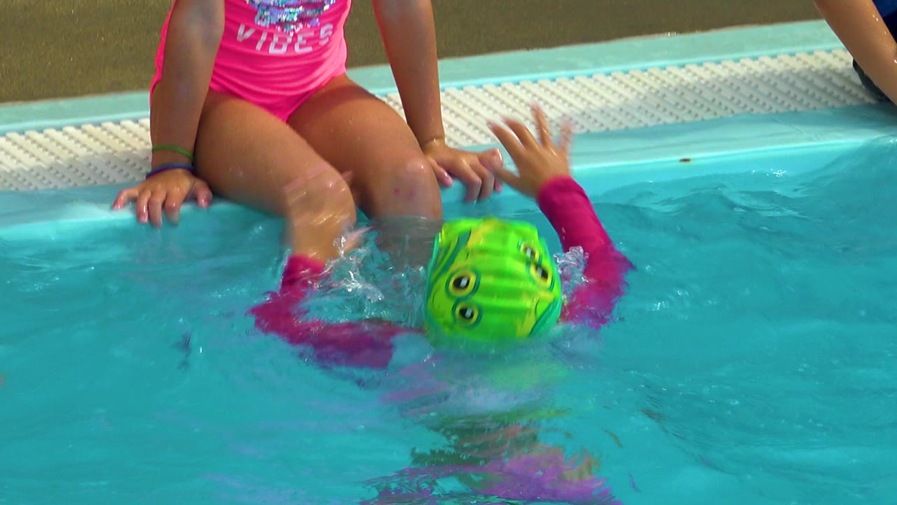 In Cabo San Lucas, Mexico, a child in a bright green swim cap swims face-down near the pool edge while a seated child in a pink swimsuit rests her hands by the water.