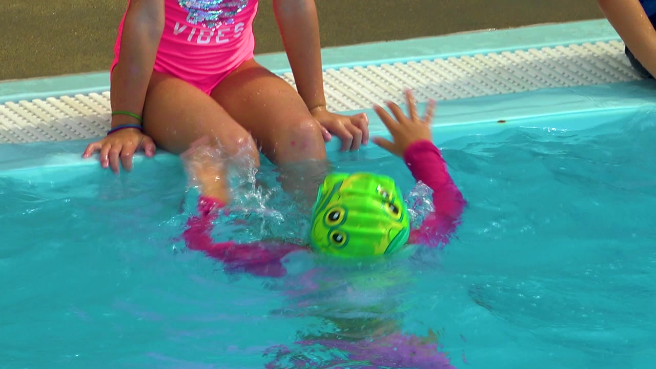 Child in green cap reaches up while swimming near the pool edge.