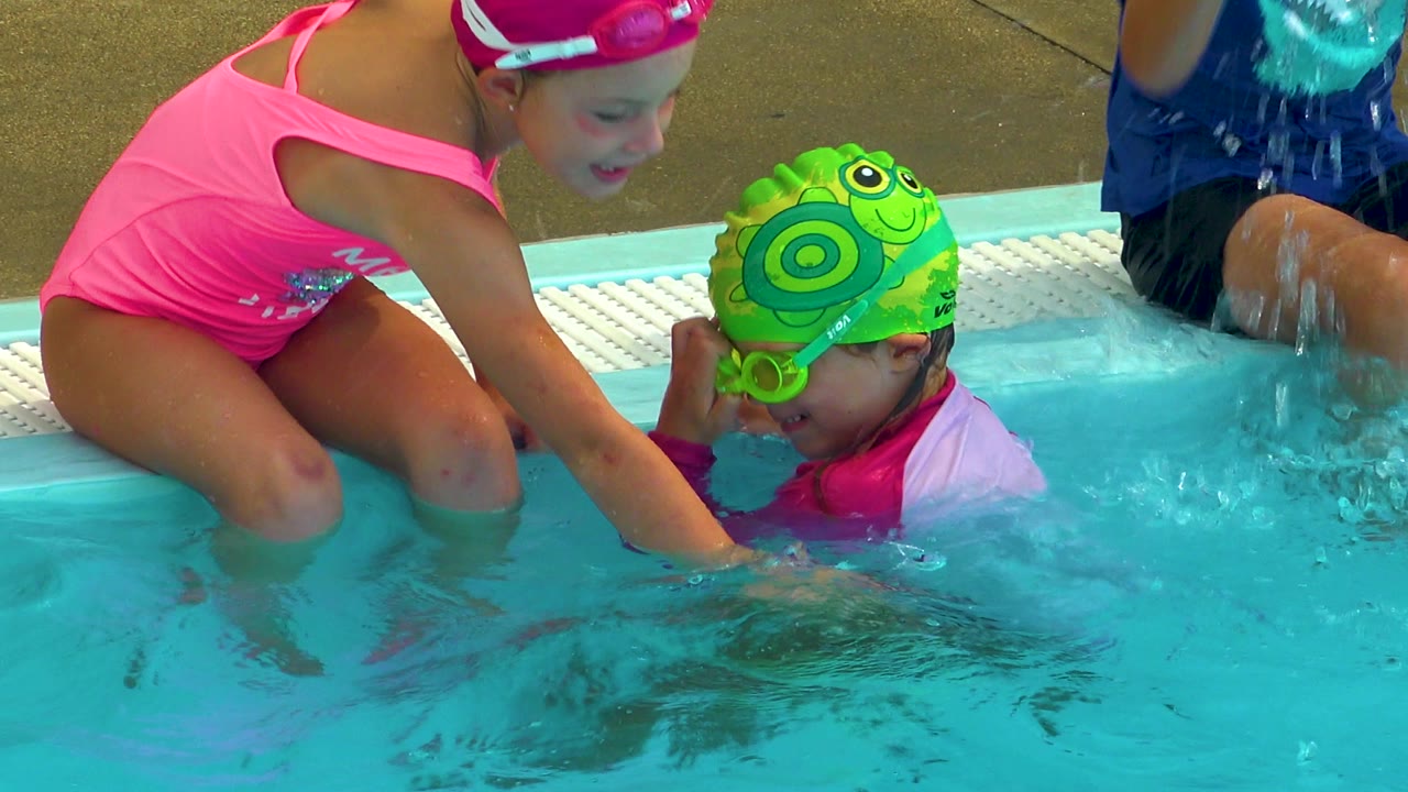At 0:00 in Cabo San Lucas, Mexico, a girl in a pink swimsuit and cap leans over the pool edge toward a younger child wearing a green turtle swim cap, with another child partly visible behind them.