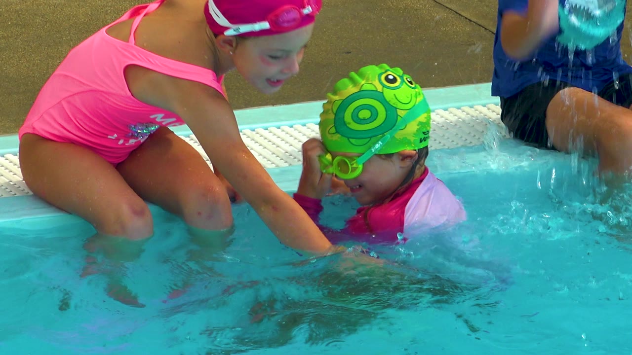 Girl in pink swimsuit reaches into the pool beside a child in a green swim cap.