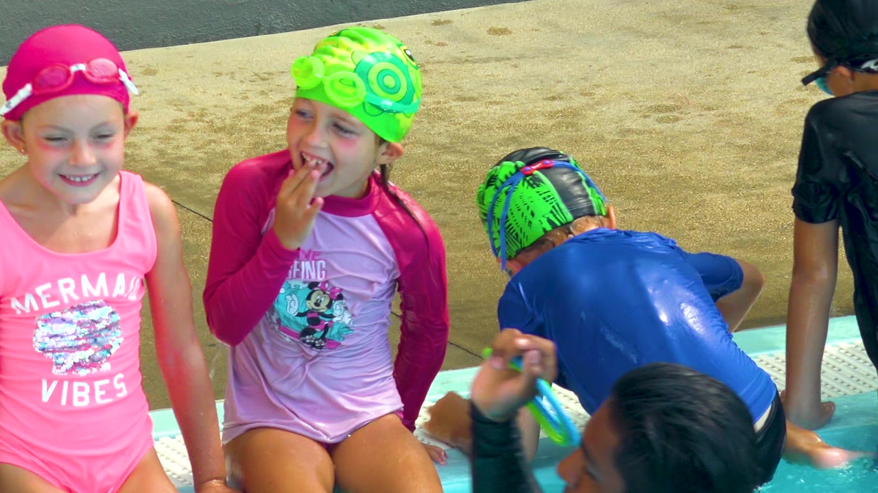 Children in Cabo San Lucas sit at a pool edge in bright swimwear, including pink and green caps, while other kids lean into the water nearby.