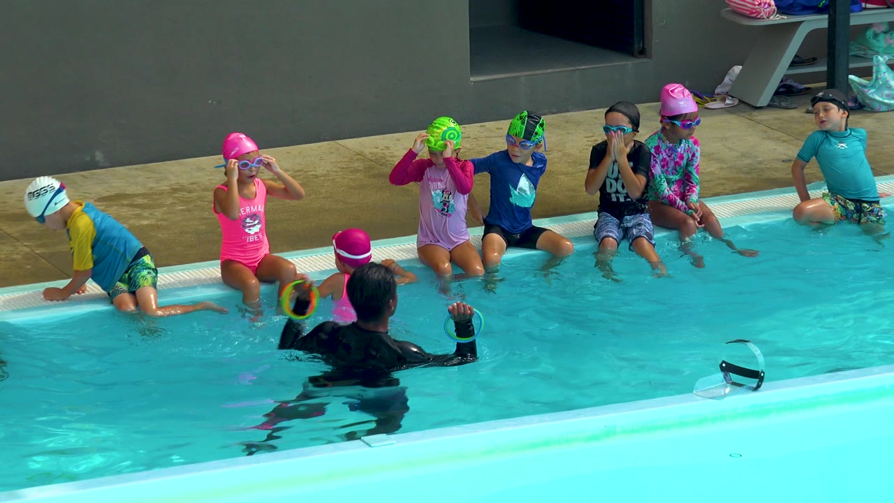 Young boys and girls in swim caps and goggles sit along a Cabo San Lucas pool edge while a coach in the foreground holds up a colorful ring.