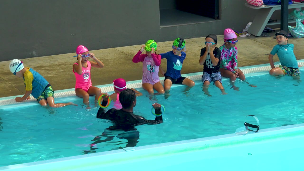 Children in swim caps watch a coach during a pool activity.