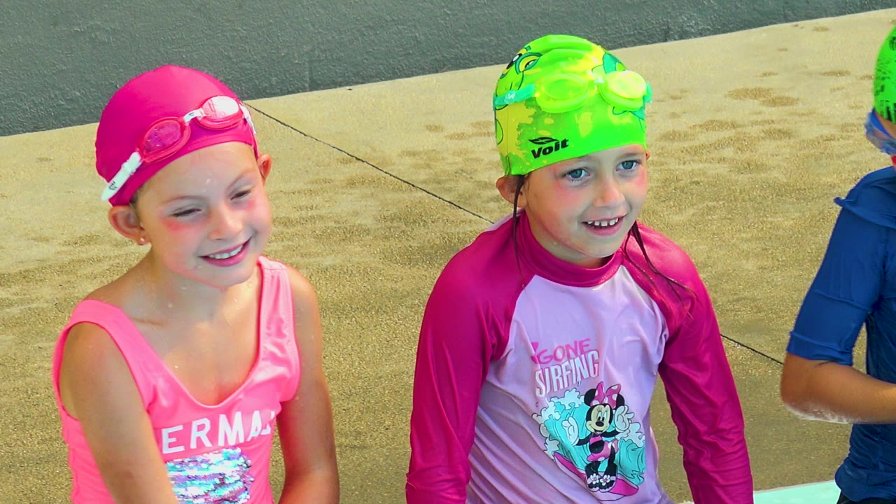 Two girls smile in swim caps beside the pool.