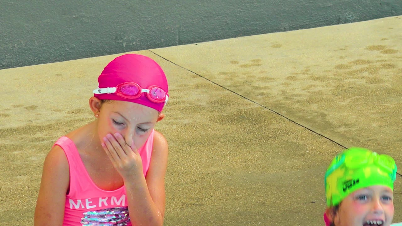 A child in a pink swim cap and goggles sits beside a pool in Cabo San Lucas, Mexico, with a second child in a green swim cap partly visible at the bottom of the frame.