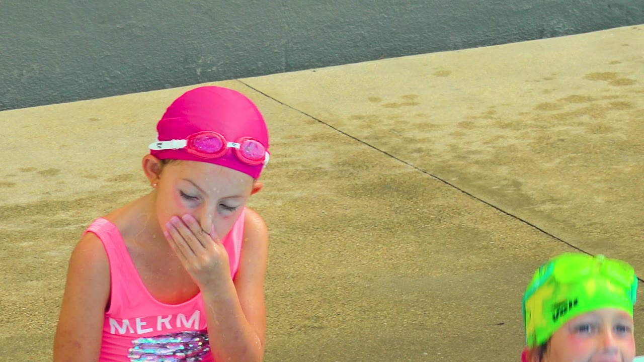 Child in pink swim cap covers their mouth while another child smiles from the pool.
