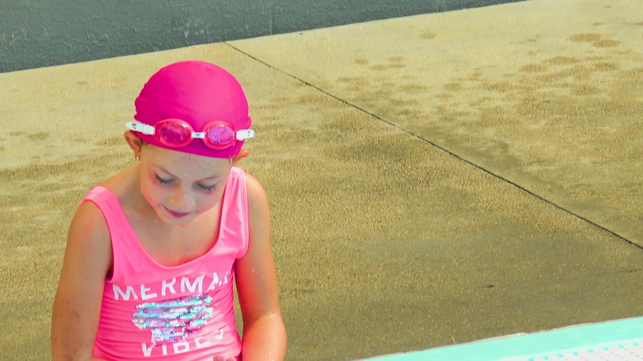 A child wearing a pink swim cap and goggles looks down near the turquoise pool edge, with another child in a green swim cap partly visible in Cabo San Lucas, Mexico.