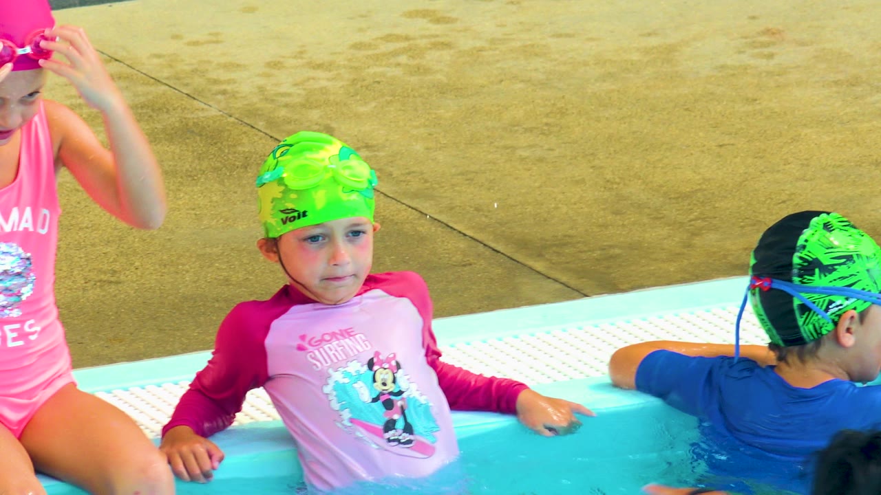 Children wait at the pool edge with goggles and swim caps.