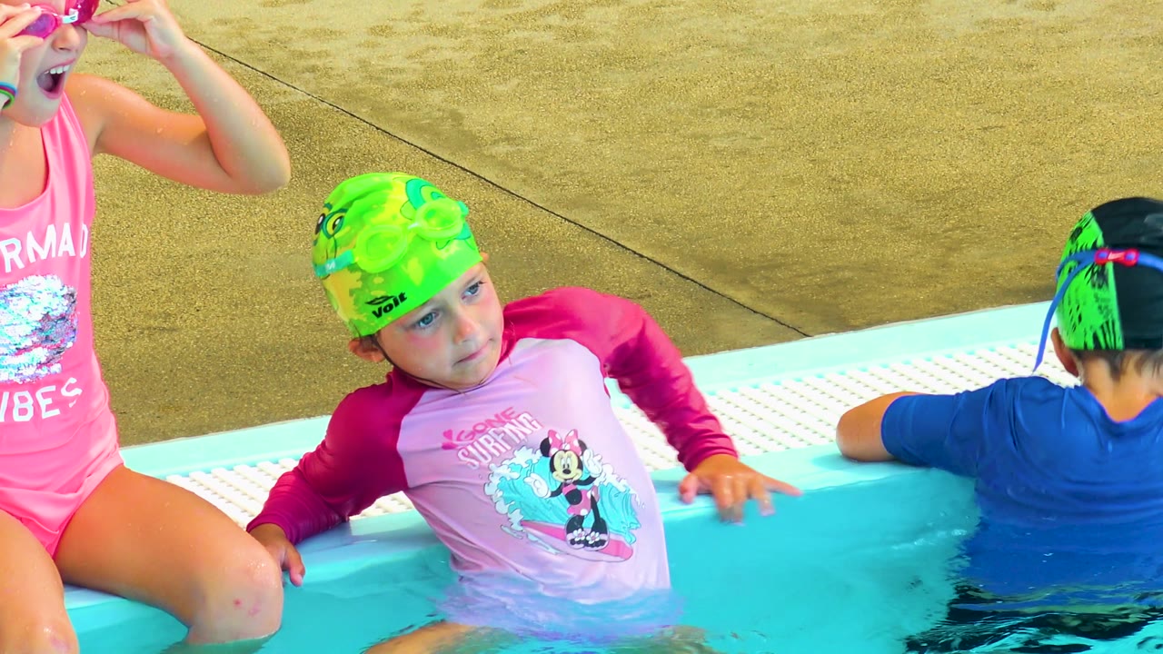 A child in a green swim cap and pink rash guard leans on the pool edge in Cabo San Lucas while another child in a blue shirt sits turned away in the water.