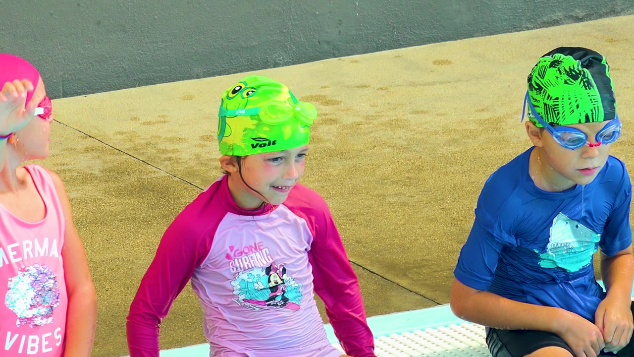 At 0:00 in Cabo San Lucas, Mexico, three children in bright swim caps and goggles sit along the pool edge, with turquoise water at the bottom of frame and concrete deck behind them.