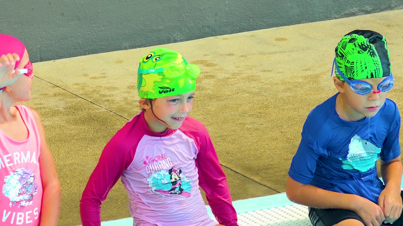 Two children in swim shirts sit beside another child in a pink swim cap.
