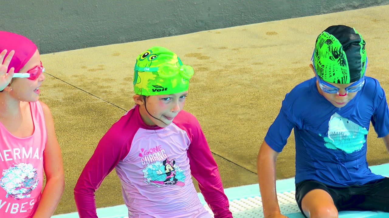 At 0:02 in Cabo San Lucas, Mexico, a child in a green cap looks toward camera while a child in a blue shirt looks down beside the pool edge.