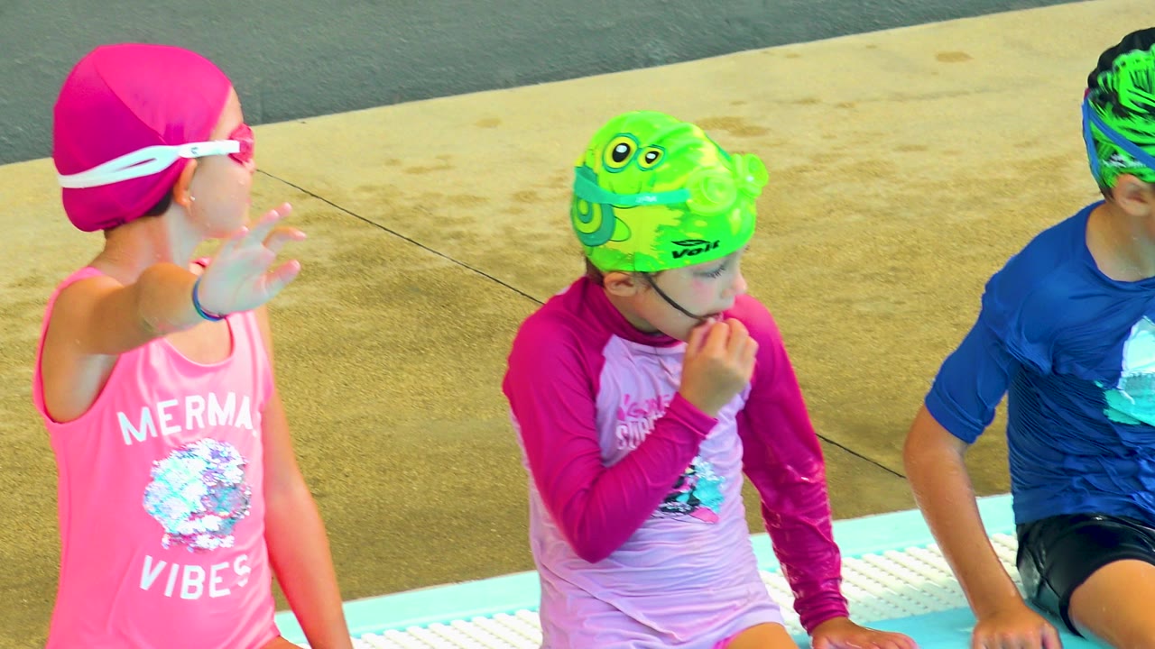 At 0:00 in Cabo San Lucas, three children in bright swim caps and goggles sit along the pool edge, with the girl in pink raising her hand near the camera.