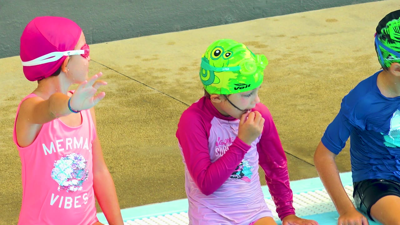 At 0:01 in Cabo San Lucas, the children remain seated on the pool edge as the child in the green swim cap looks down and touches their face.