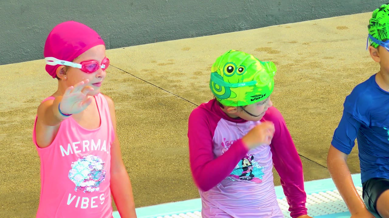 At 0:02 in Cabo San Lucas, the girl in the pink cap and goggles turns more toward the camera while two other children sit beside her on the pool edge.