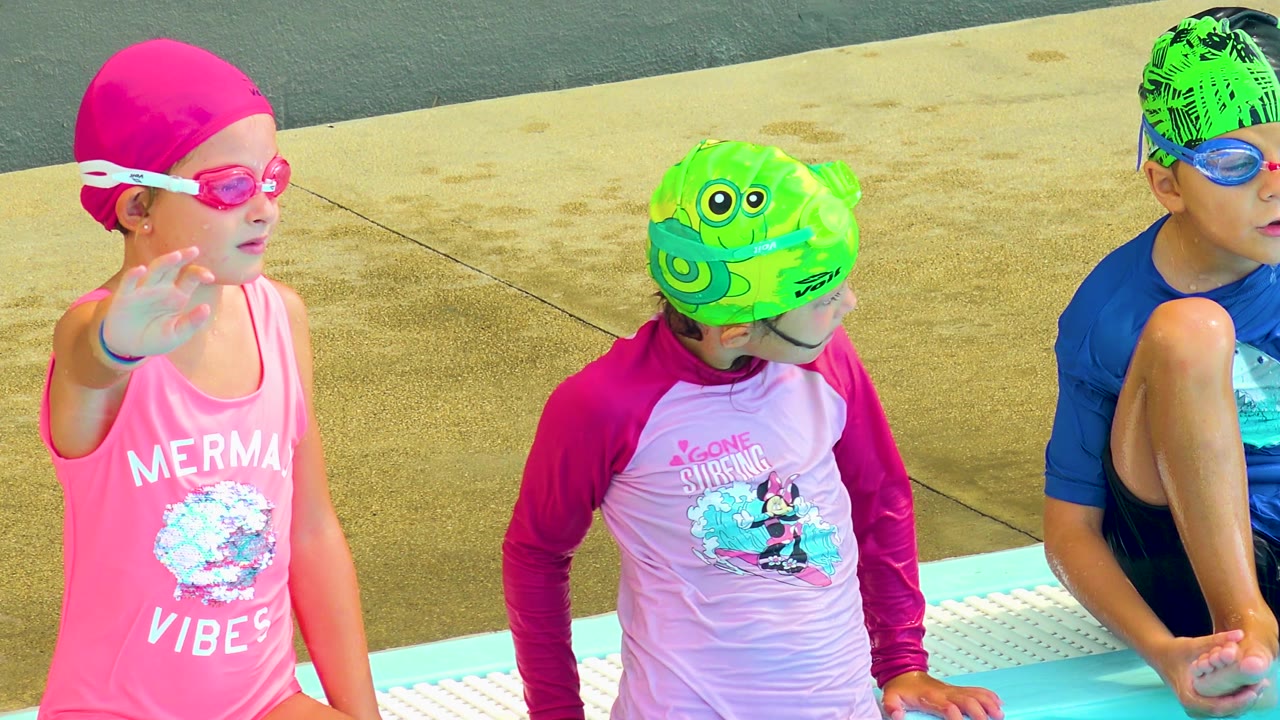 Three children in swim gear sit together at the pool edge.