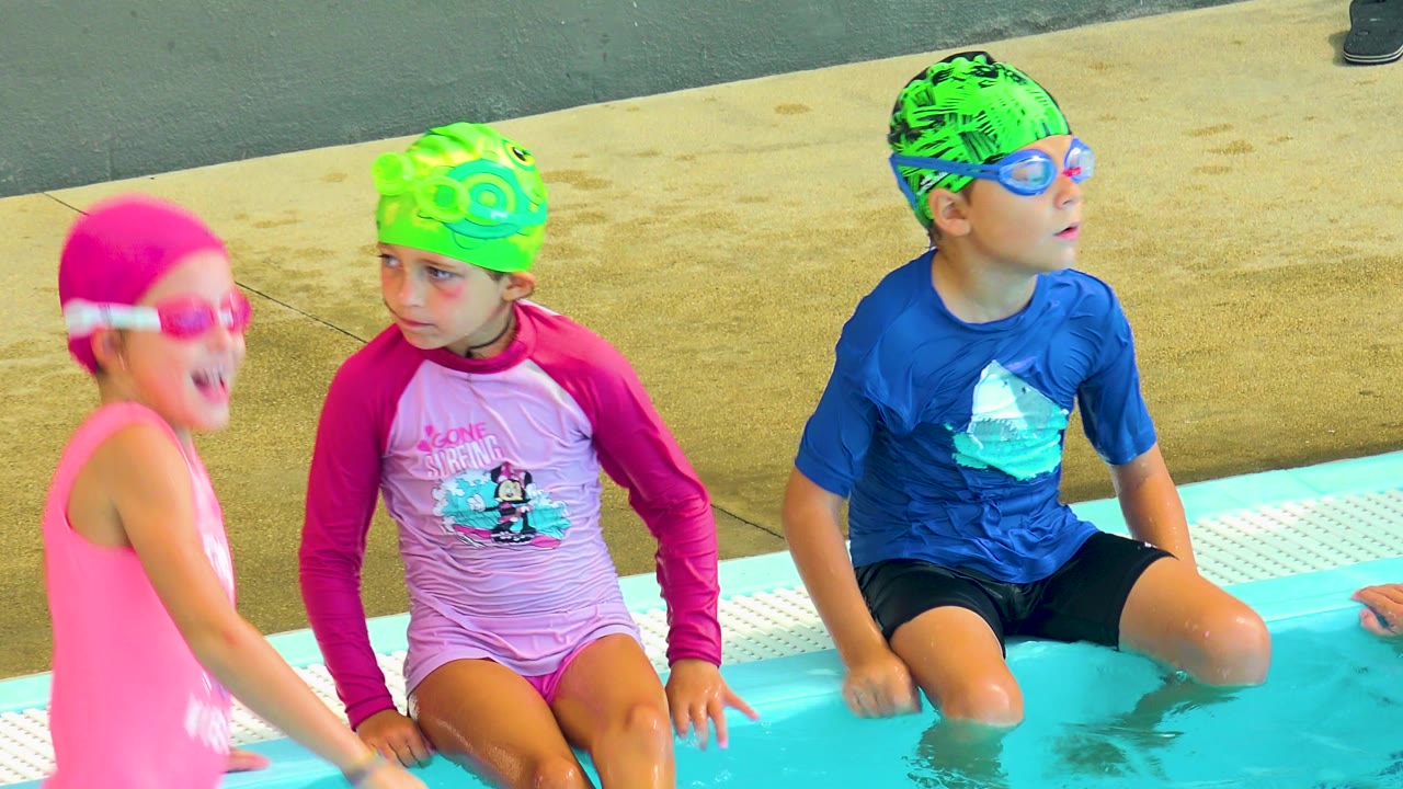 Three children sit and stand at the edge of an outdoor pool in Cabo San Lucas, Mexico, including a girl in a pink cap, a child in a green cap, and a boy in a blue swim shirt.
