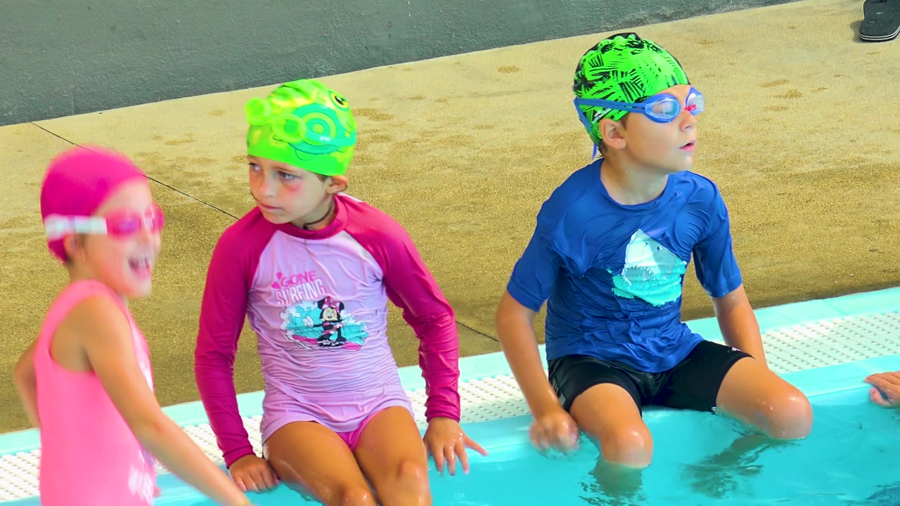 Children in bright swim gear waiting beside pool water.