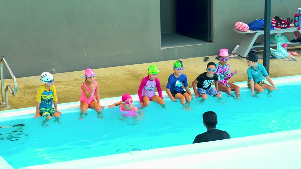 In Cabo San Lucas, Mexico, several children in swim caps and goggles sit along the pool edge as an adult in dark clothing faces them from the foreground water.