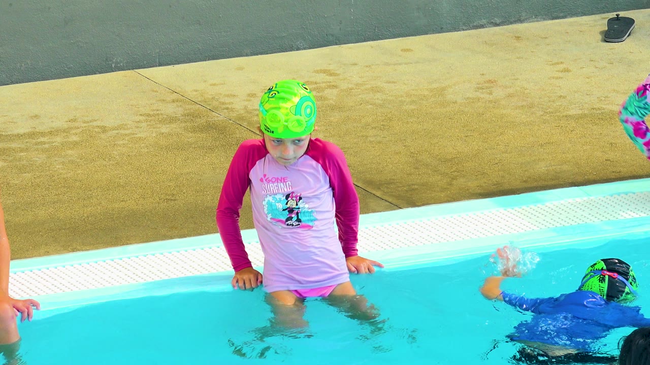 In Cabo San Lucas, Mexico, a child in a green swim cap and pink swim shirt rests hands on the pool edge while another swimmer is partly visible in the water.