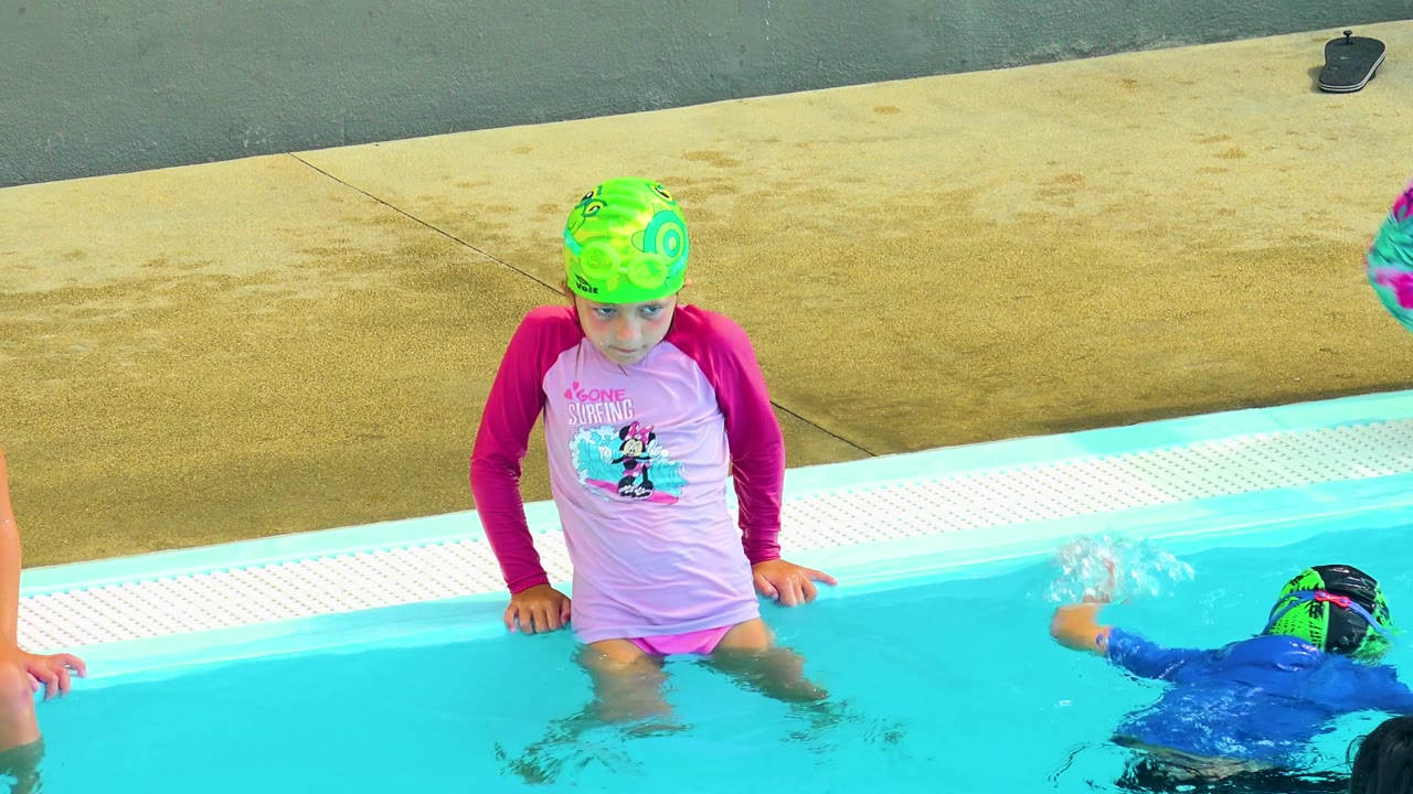 Child at the pool edge while a swimmer surfaces nearby.