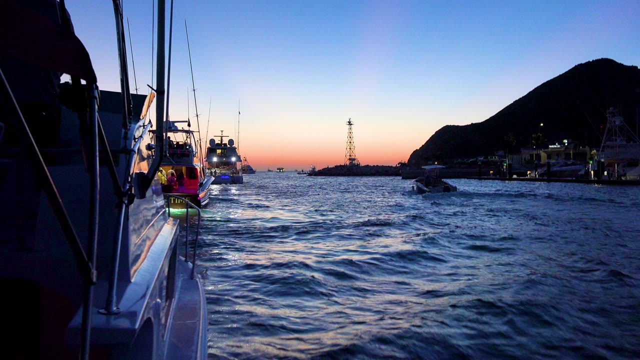 Cabo San Lucas boats travel over choppy blue water beside a yacht railing in the left foreground, with a dark hillside and warm horizon light, no visible people.