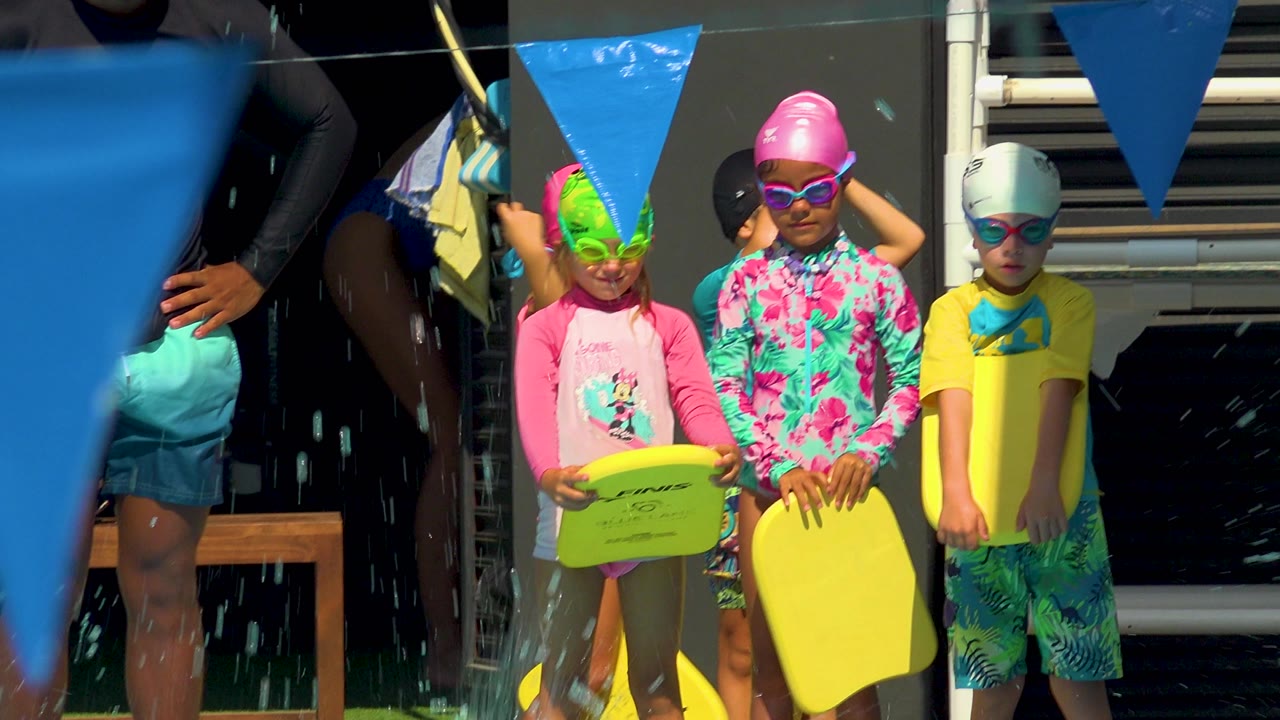 At a Cabo San Lucas pool, three small children in bright swim caps and goggles stand near the water with yellow kickboards while blue swim flags hang overhead.