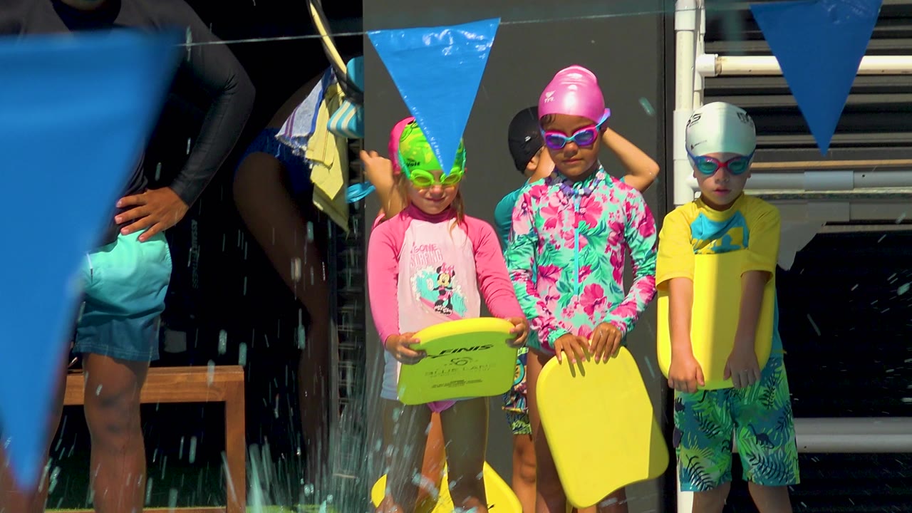 Children wait at the pool edge with kickboards and swim flags overhead.