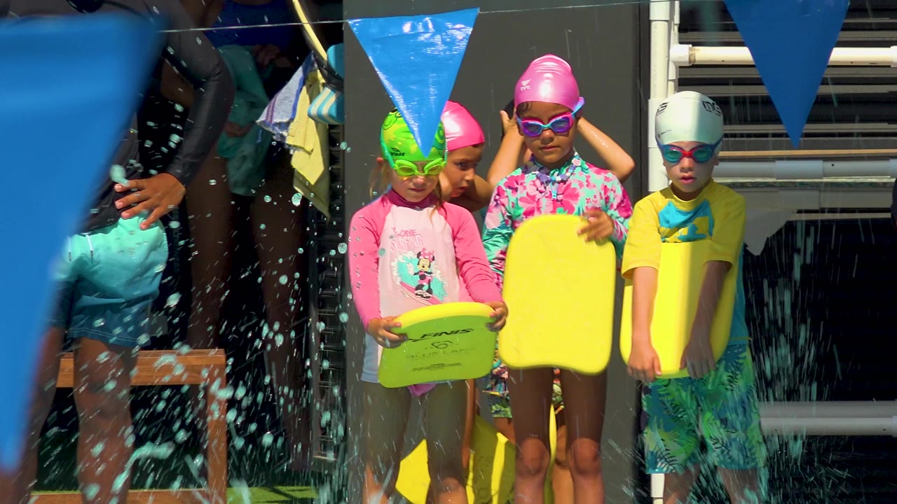 Water splashes across the foreground as several children in swim caps and goggles stand at the Cabo San Lucas pool edge with yellow kickboards and blue flags above them.