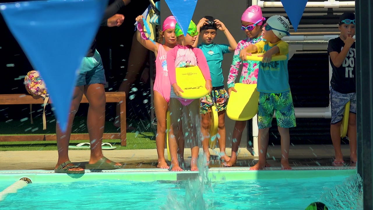 Children in swim caps and goggles stand at a turquoise pool edge in Cabo San Lucas, Mexico, with yellow kickboards, blue pennants, and an adult instructor partly visible.