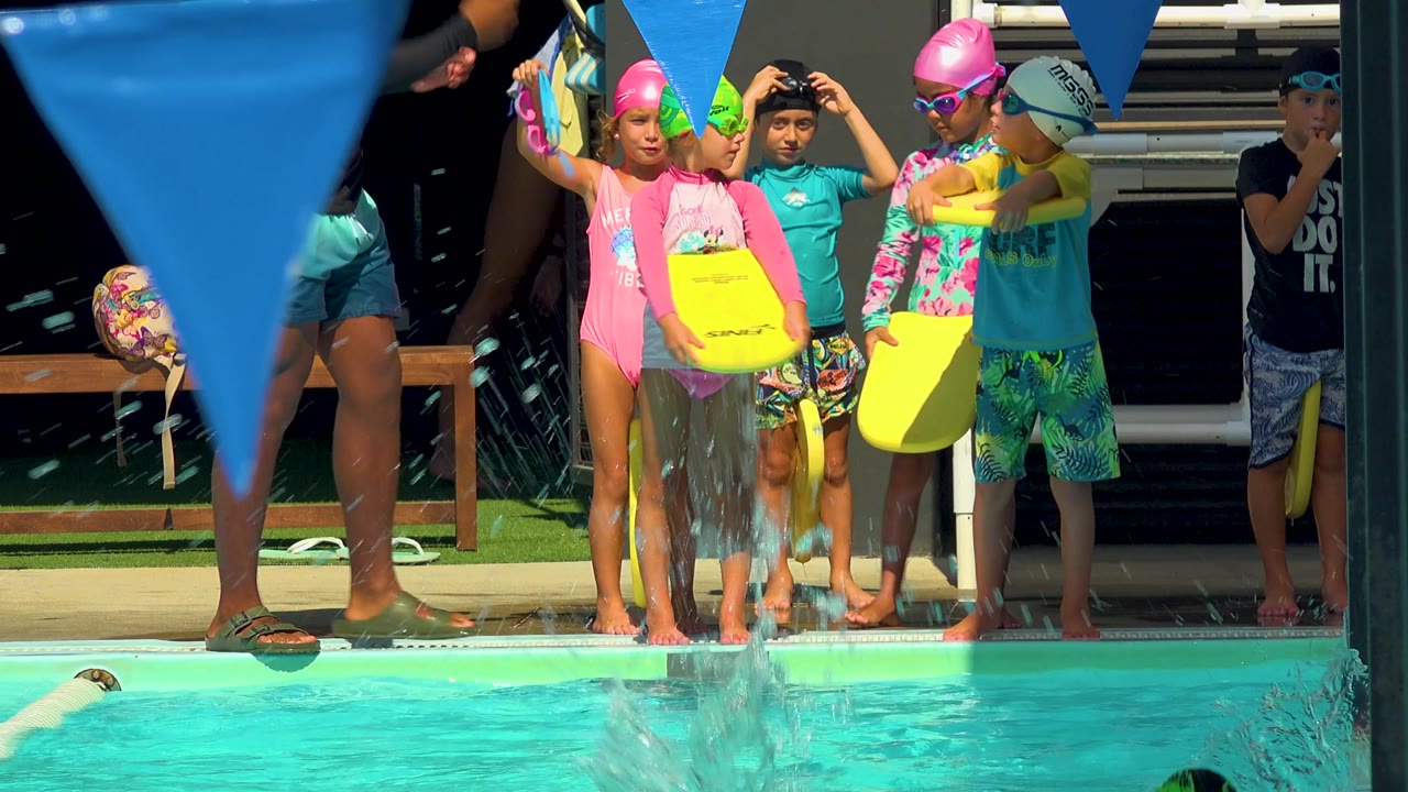 Several children in bright swimwear wait beside the pool in Cabo San Lucas, Mexico, as water splashes near the edge and blue triangular pennants hang above.