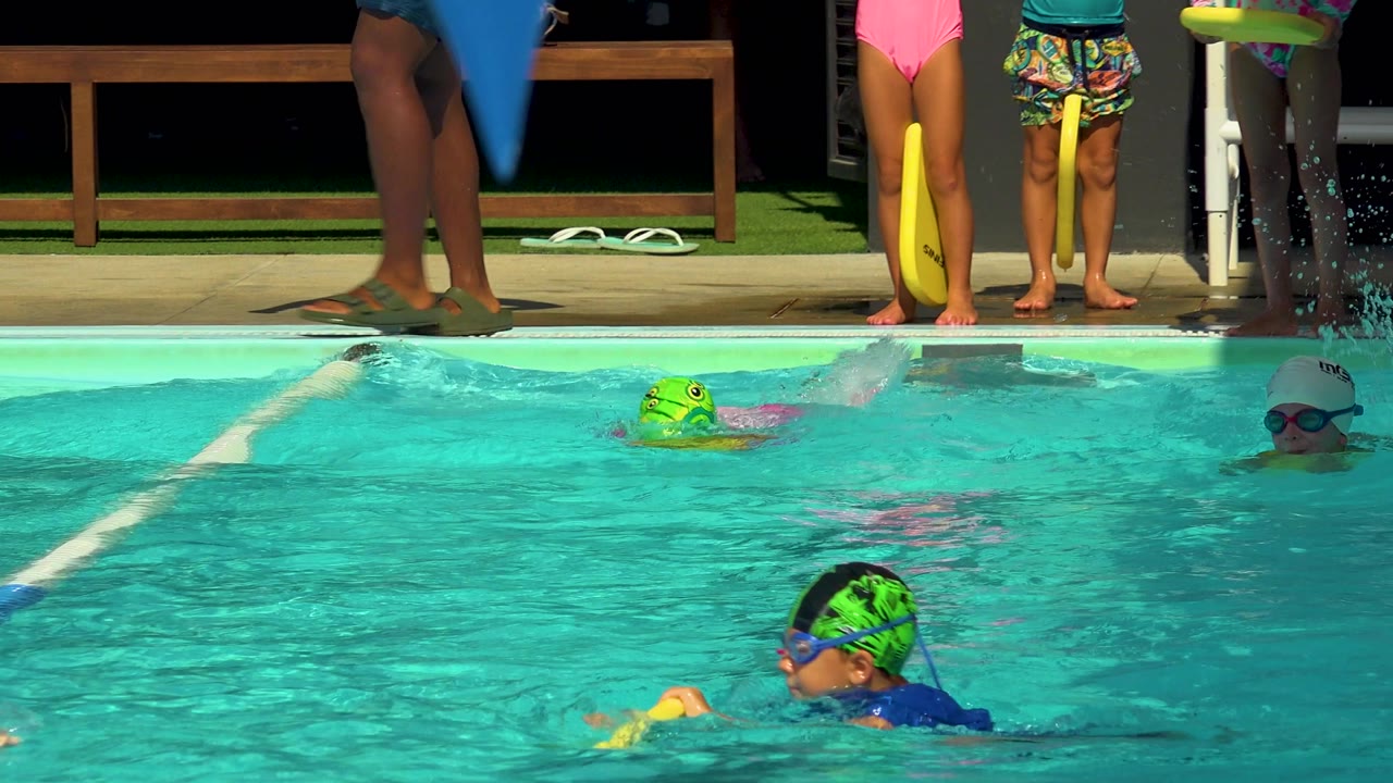 At 0:02 in Cabo San Lucas, one child swims near the right with a yellow kickboard while another child in a green cap moves farther back in the lane.