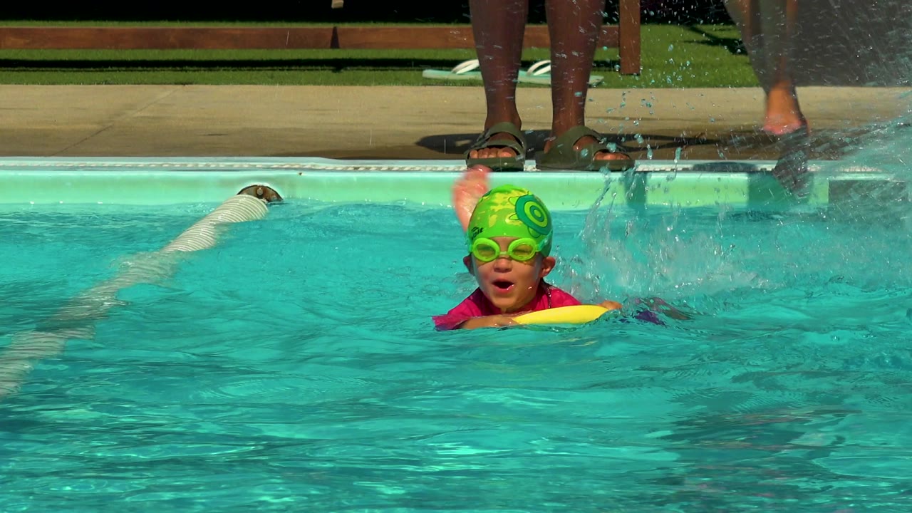 At 0:00 in Cabo San Lucas, Mexico, a young boy wearing green goggles and a green swim cap holds a yellow flotation noodle in turquoise pool water as splashes rise near the deck.