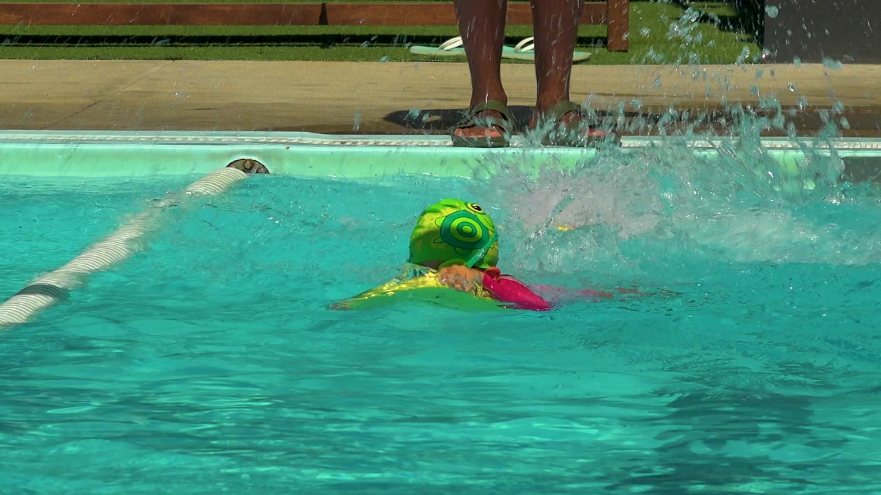 At 0:02 in Cabo San Lucas, Mexico, the green-capped child turns away from camera with a yellow float under his arms as a burst of pool spray fills the right side of the frame.