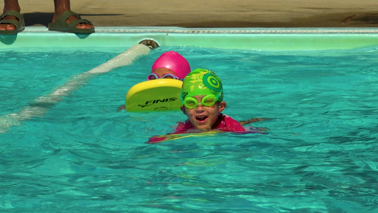 At 0:00 in Cabo San Lucas, Mexico, children in bright swim caps and goggles float near yellow kickboards in turquoise pool water, with adult feet visible on the pool deck.