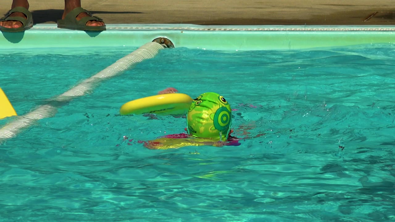At 0:02 in Cabo San Lucas, Mexico, a child in a green swim cap swims away from the camera beside a white lane rope as a yellow kickboard tilts above the water.