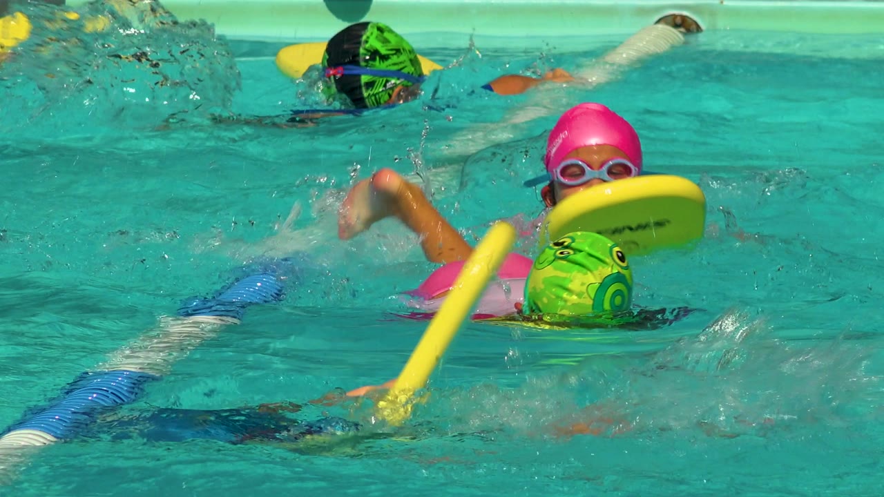 Two children move forward in pool lanes with kickboards and swim caps.