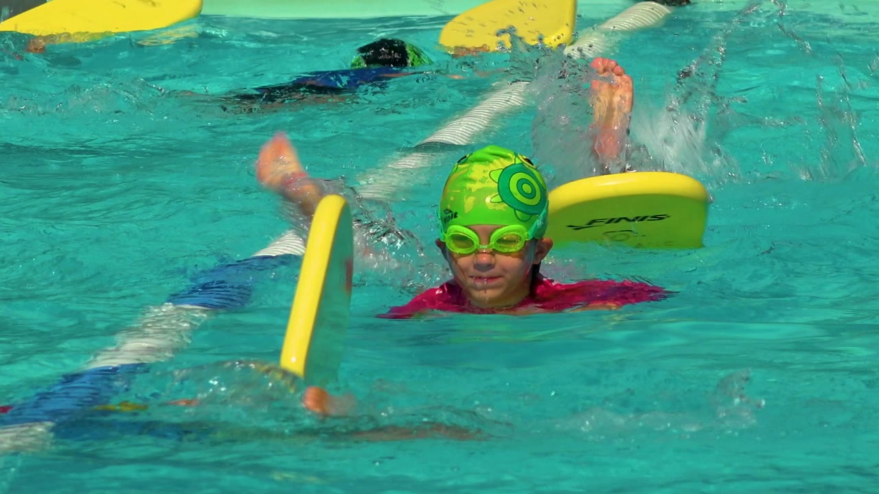 A child in a green patterned swim cap and goggles faces the camera beside a yellow kickboard, with other swimmers and lane dividers in the Cabo San Lucas pool.