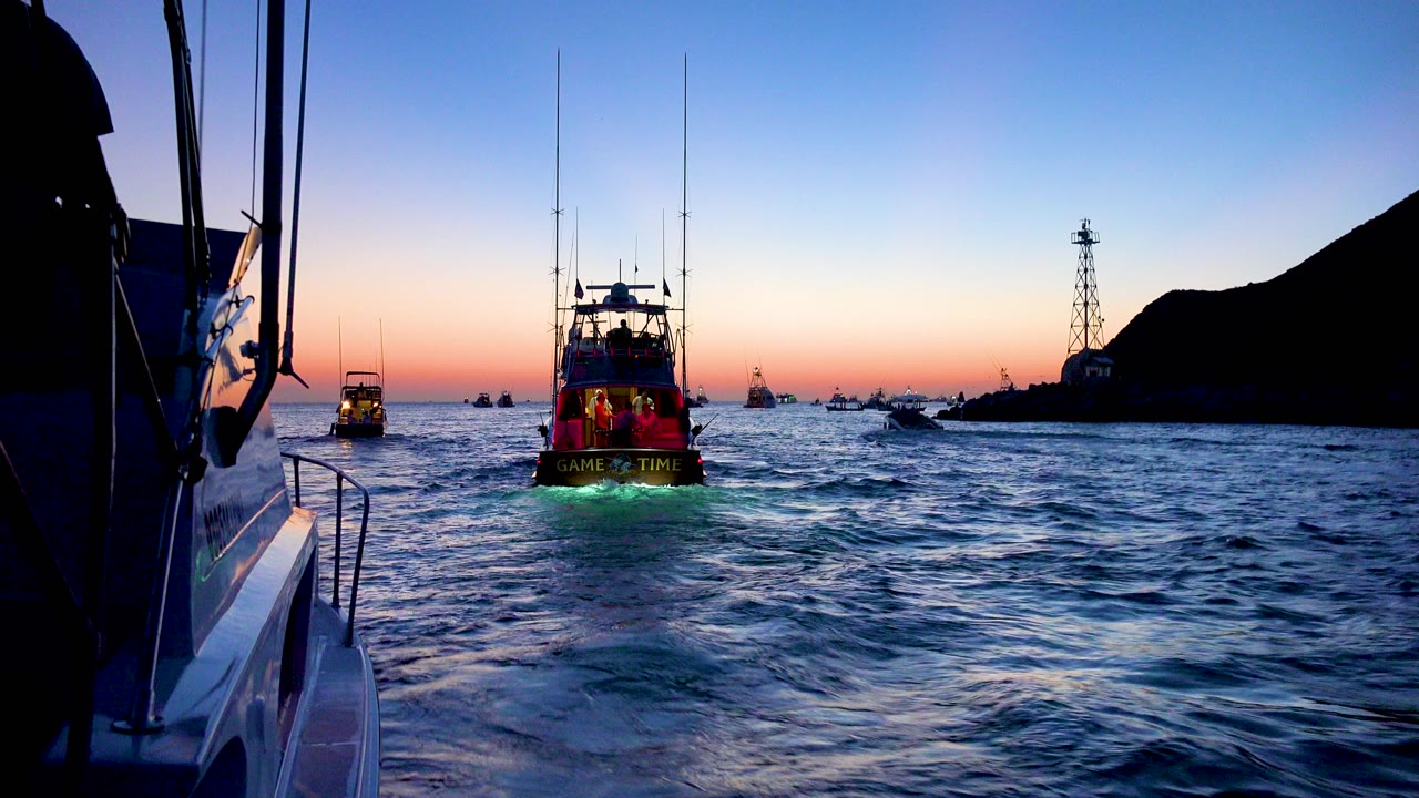 Cabo San Lucas dusk ocean view from a boat, with a neon-lit boat centered ahead, a left-side rail in foreground, distant vessels, dark shore silhouettes, and no visible people.