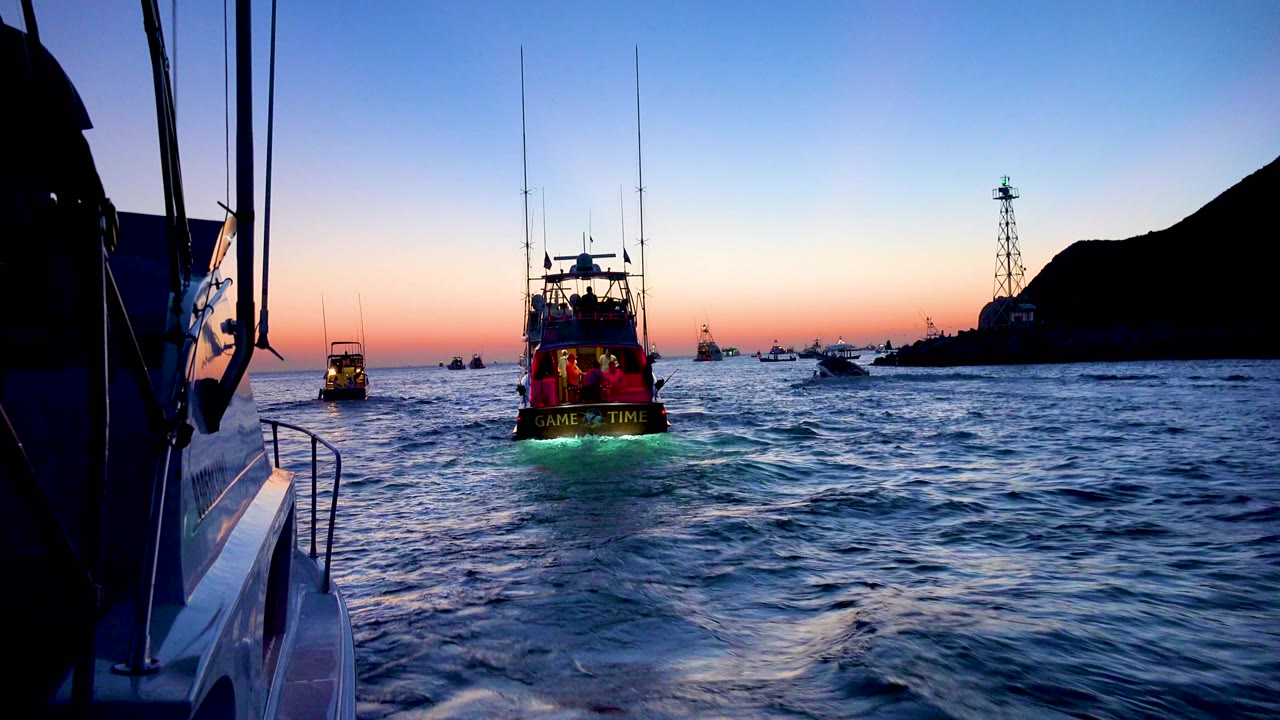 Cabo San Lucas evening boat view with a central vessel showing green stern illumination, blue water in the foreground, an orange horizon line, silhouetted land at right, and no visible people.