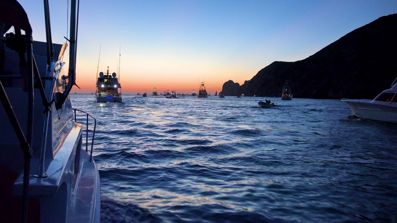 Cabo San Lucas ocean view at sunset with a lit vessel near center, small boats on the horizon, dark cliffs to the right, and boat railings framing the foreground.