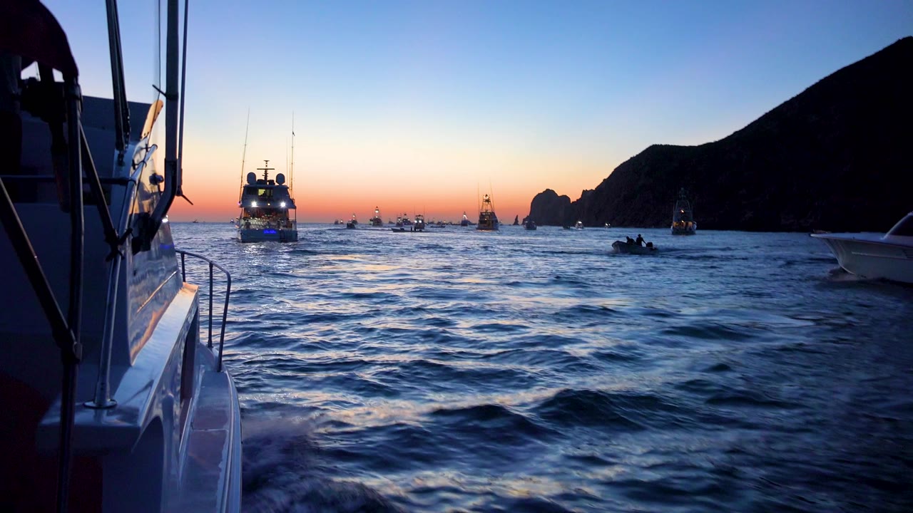 Cabo San Lucas sunset water view with the camera looking past a boat edge toward a central lit vessel, multiple small boats, and dark coastal rock silhouettes.