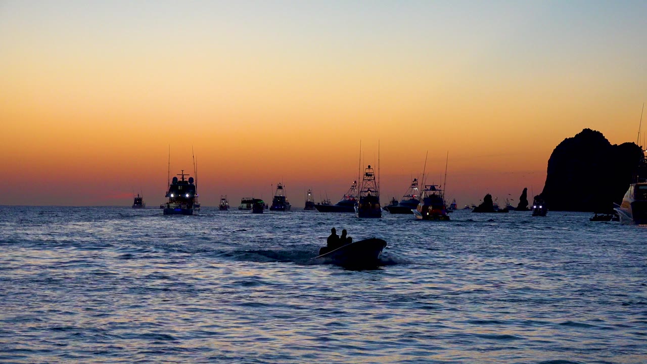 At 0:00 in Cabo San Lucas, a small silhouetted boat moves across rippled water while larger boats and a tall rock formation sit against a yellow and red dusk sky.