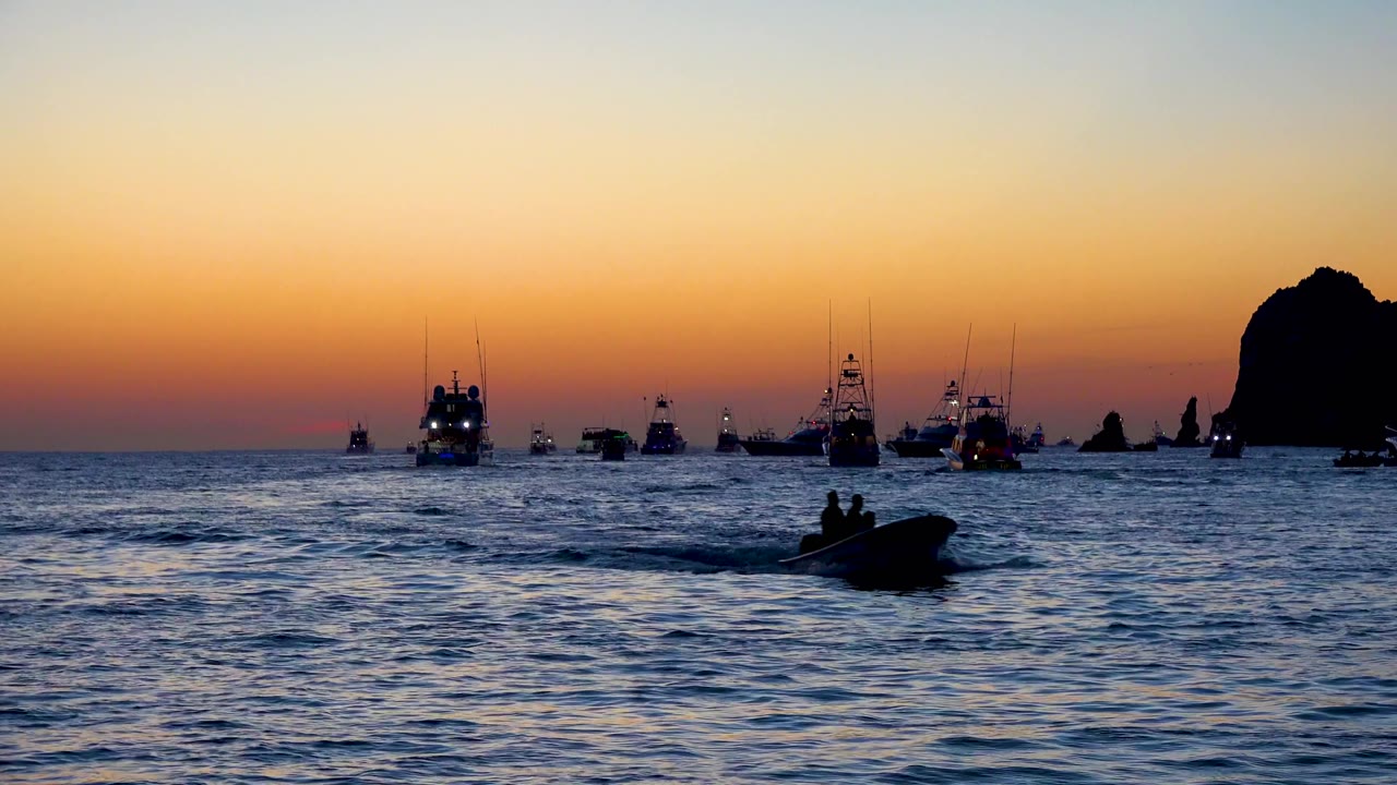 At 0:02 in Cabo San Lucas, the foreground boat remains a dark silhouette over blue rippled water, with multiple distant boats lined under the orange dusk band.