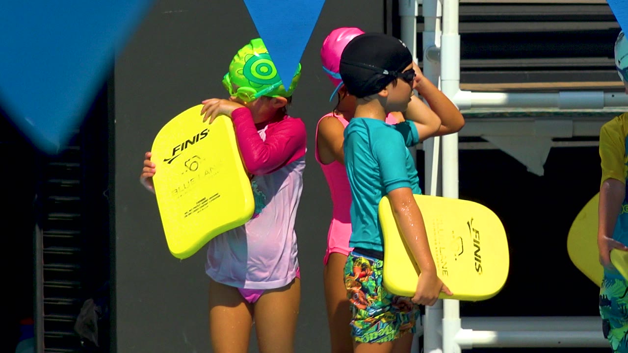 At a Cabo San Lucas poolside, children in bright swim caps and goggles stand close together while holding yellow FINIS kickboards under strong sun.
